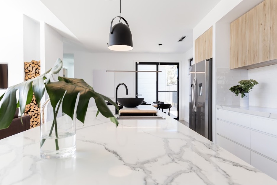 Modern kitchen featuring a sleek marble island with a vase of green leaves, black pendant light, wooden cabinets, and stainless steel fridge. Bright and airy.