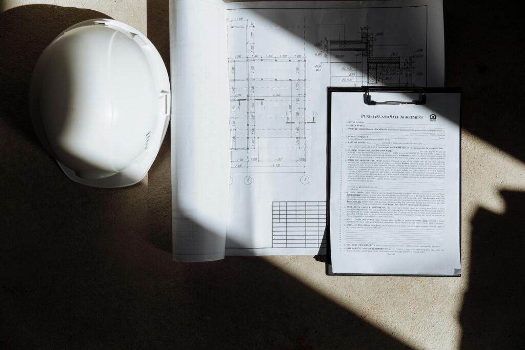 A white hard hat sits next to architectural blueprints and a document on a clipboard, lit by sunlight casting shadows, conveying a professional tone.