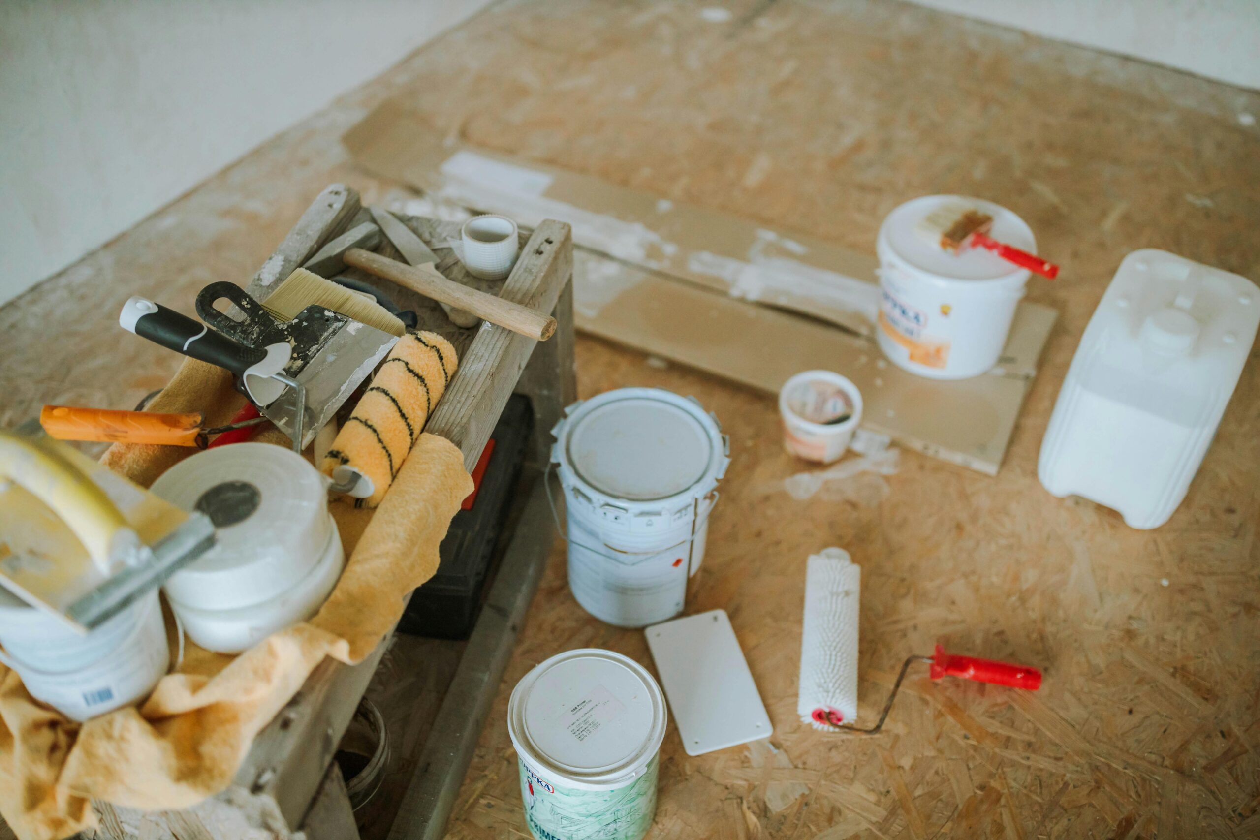 Painting supplies on a wooden floor; includes rollers, brushes, paint cans, and a toolbox, suggesting a home renovation scene in progress.