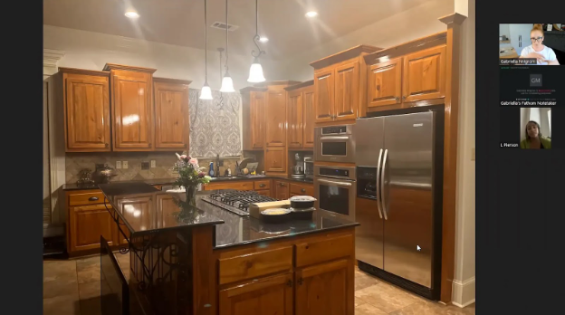 A warm kitchen with wooden cabinets and stainless steel appliances, featuring a central island with a bouquet of flowers. Two video call participants are visible in the top right.