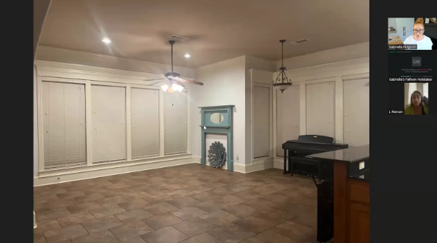 A spacious, dimly lit room with brown tile flooring and large windows covered by blinds. There's a ceiling fan, an old fireplace, and a piano. An online meeting with two visible participants is in progress on the right side.