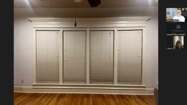 A well-lit room with four tall windows, white blinds fully closed, and beige walls. Hardwood floor adds warmth. Two small video call thumbnails in the image corner.
