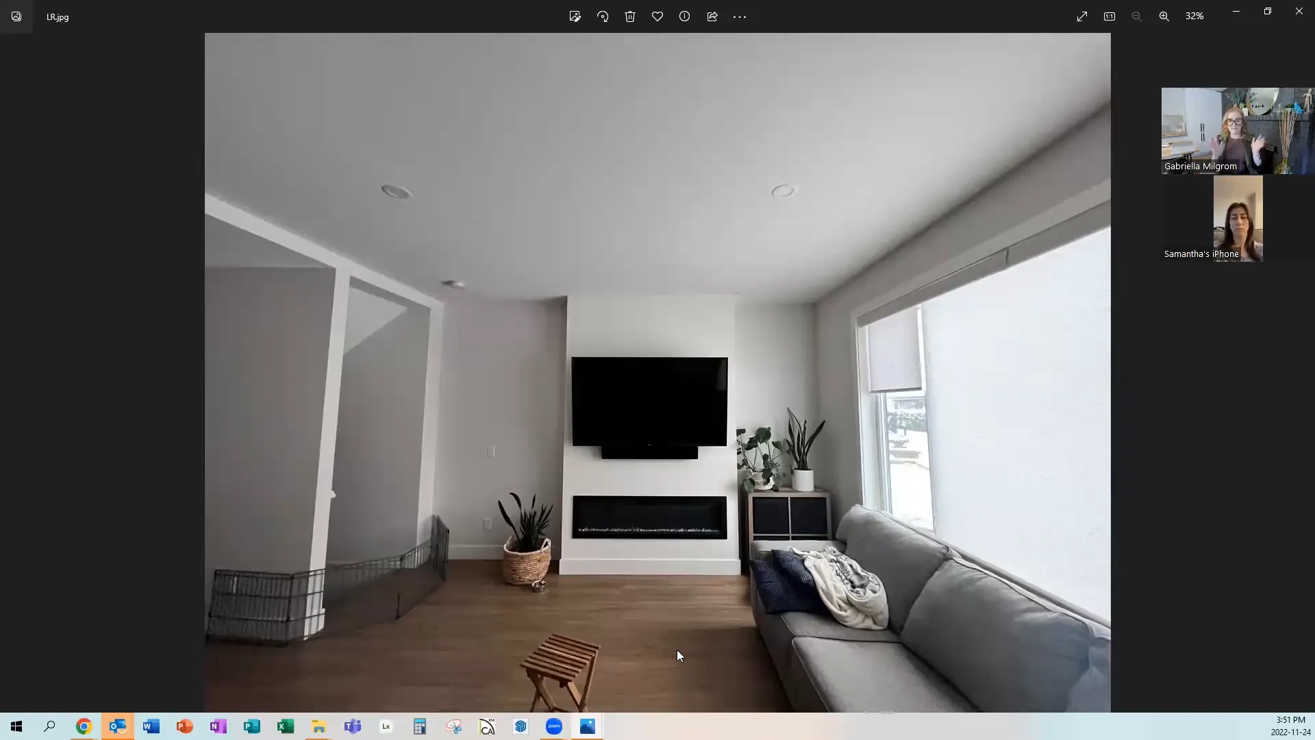 Modern living room with a wall-mounted TV above a linear fireplace. Gray sofa on the right, a small wooden stool, and potted plants add decor. Natural light streams through large windows, creating a calm, cozy atmosphere.