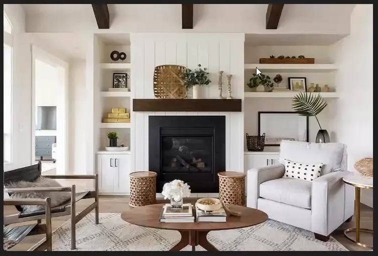 A cozy living room features a white fireplace with dark wood accents, surrounded by shelves holding books and decor. A plush armchair, modern coffee table, and geometric rug enhance the warm, inviting atmosphere.