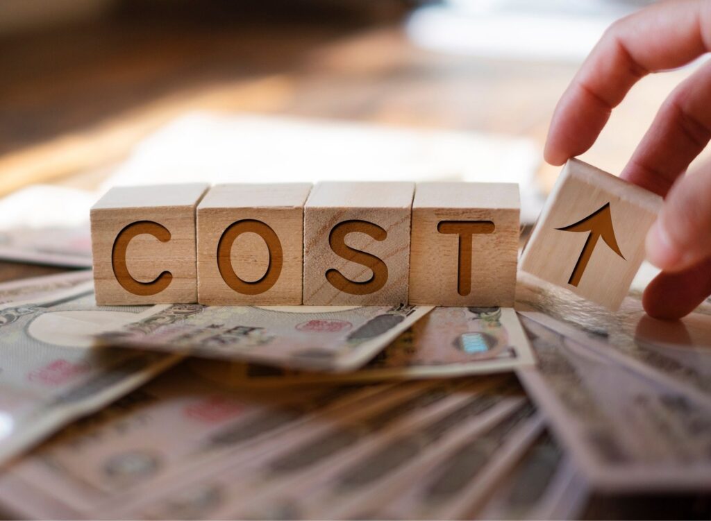 Wooden blocks spell "COST" on a bed of currency notes, with a hand adding an arrow block indicating a rise, suggesting increasing expenses.