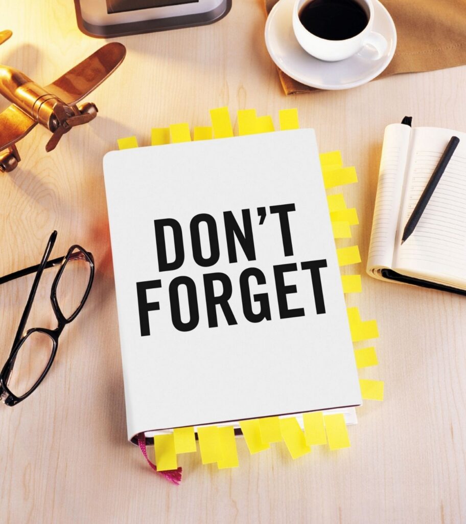 Notebook with "DON'T FORGET" text, surrounded by yellow sticky notes on a desk. Nearby are glasses, a model airplane, an open notebook, and a coffee cup.