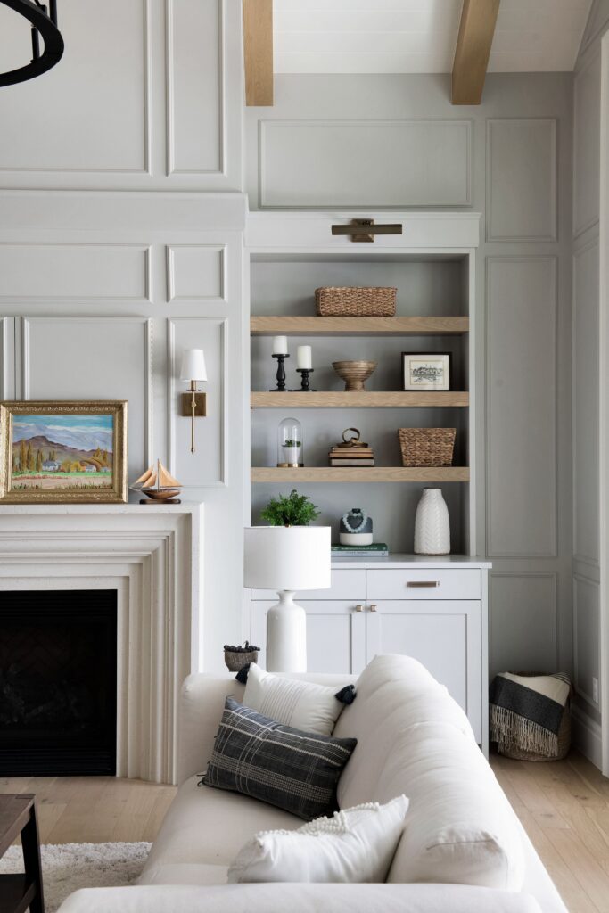 Elegant coastal cottage living room with custom white built-ins, natural oak floating shelves, decorative wall panel moulding, exposed wood ceiling beams, a white fireplace surround, and neutral layered furnishings creating a warm, timeless interior.