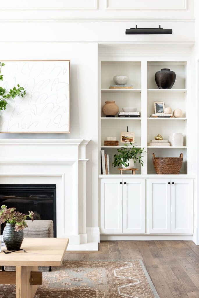Bright minimal traditional living room with custom white built-in bookshelves, lower storage cabinets, a classic fireplace mantel, neutral décor styling, warm wood flooring, and layered textures for a timeless, elevated design.
