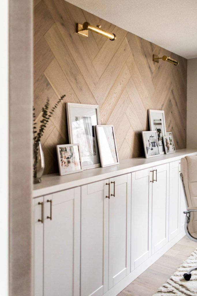 Modern home office with a white built-in storage wall, custom herringbone wood accent wall, brass picture lights, shaker-style cabinetry, and neutral décor creating a warm and functional workspace.