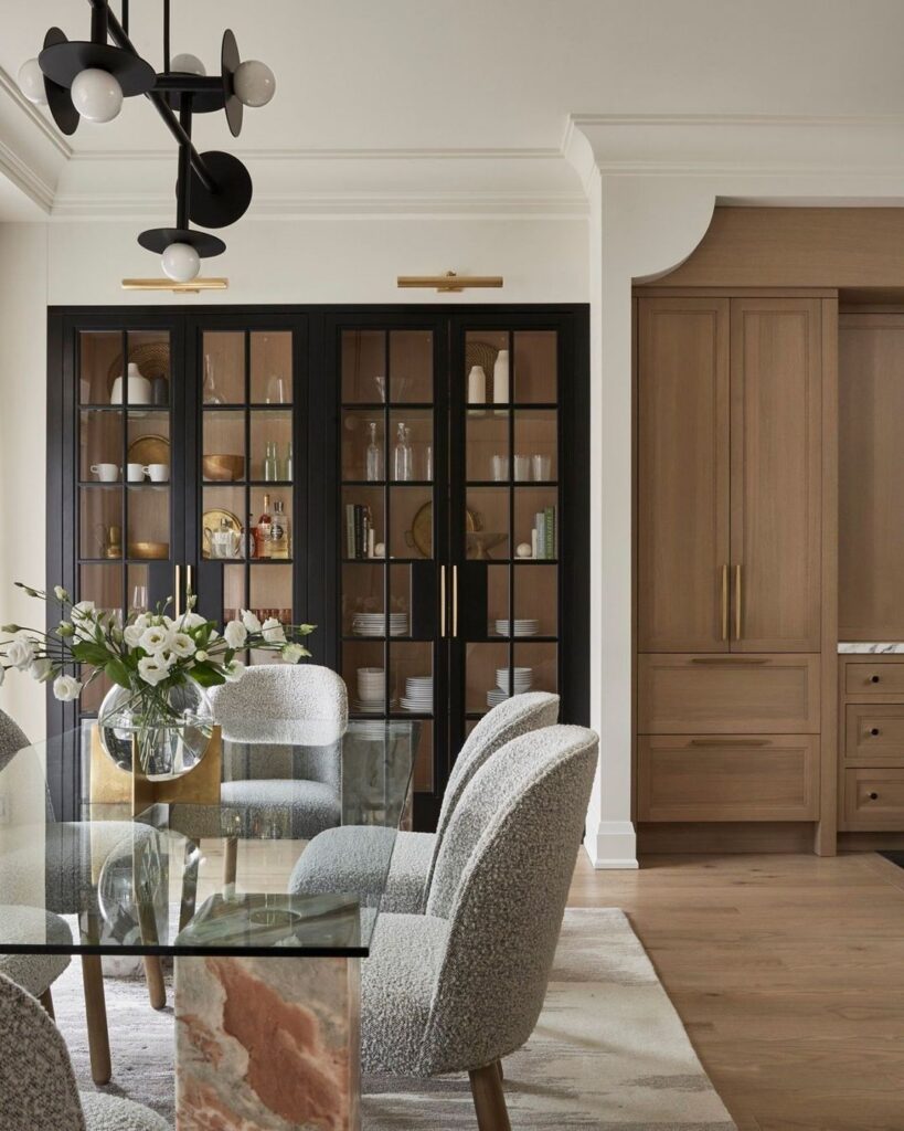 Modern traditional dining room with a black glass-front china cabinet, white walls with crown moulding, warm oak cabinetry, boucle dining chairs, a glass dining table, and layered black and brass accents.