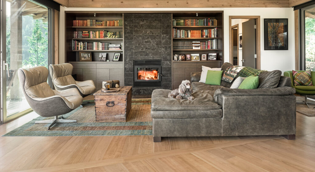 A cozy living room features a stone fireplace with a warm fire, surrounded by bookshelves. A leather sofa with a dog, modern chairs, and a wooden coffee table enhance the inviting ambiance.