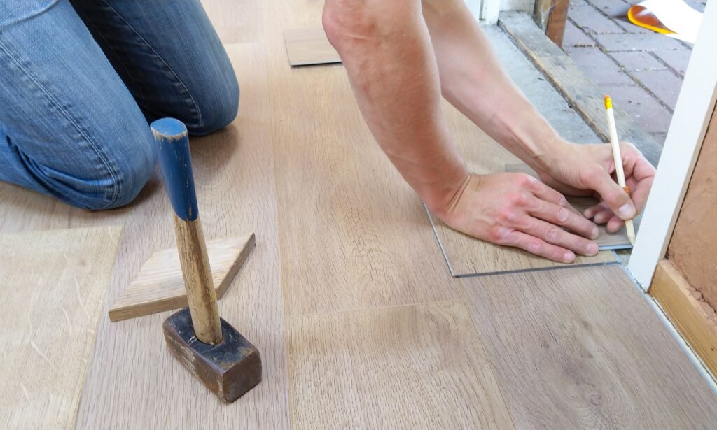 A person kneels on a wooden floor, marking a tile with a pencil next to a measuring tool. A hammer rests nearby, conveying focus and precision in renovation work.